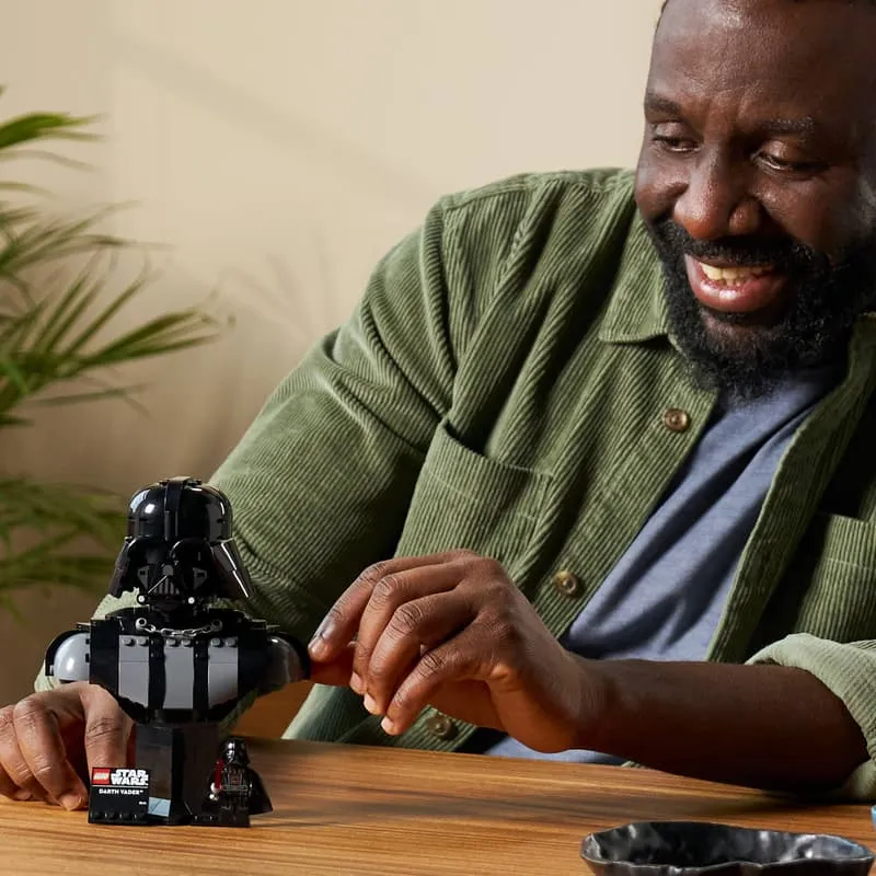 A man smiling happily as he interacts with the assembled Lego Darth Vader Bust 75439 on a wooden table, emphasizing the enjoyment of the building and display experience.