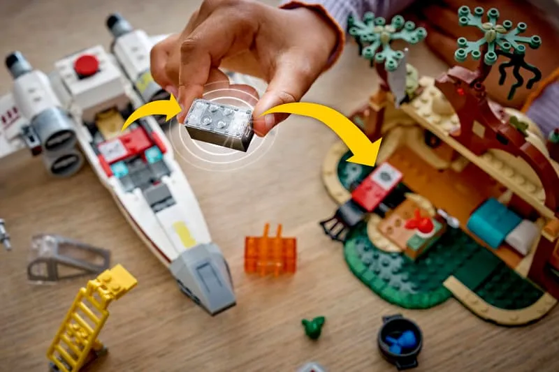 A hand transferring a black Lego Smart Brick from an X-wing (not part of this set) to a designated slot in the Lego SMART Play™: Yoda's Hut and Jedi Training 75422 set.