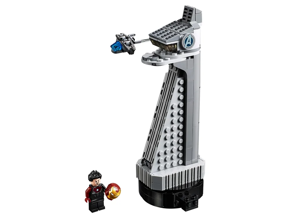A LEGO micro-build of the Avengers Tower on a black base, featuring a grey tapering structure, a detailed top platform with an Avengers logo, a small flying vehicle, and a Tony Stark minifigure holding an Iron Man helmet. The background is white.