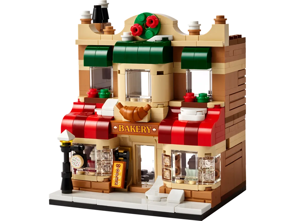 A detailed front view of the LEGO 40696 Bakery building, featuring a tan and dark tan facade, a red and white striped awning over the entrance, and green awnings over the upper windows. A golden croissant element is positioned above the "BAKERY" sign, and a festive red and green wreath adorns the rooftop. A streetlamp and an "OPEN" sign are visible on the sidewalk.