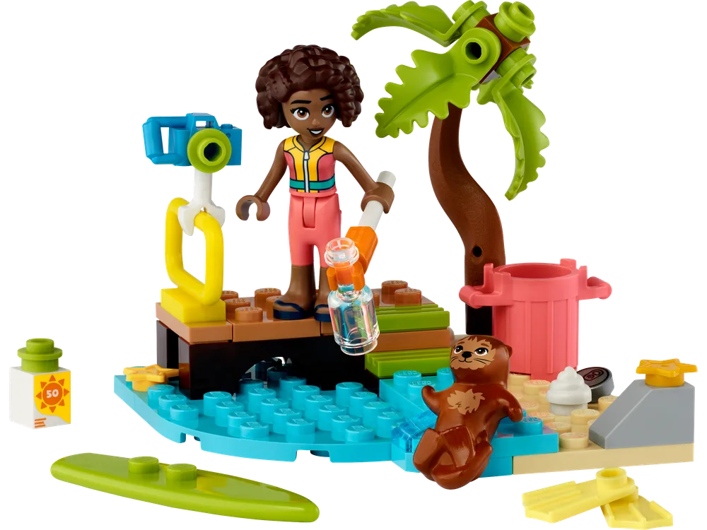A LEGO Friends minifigure with a grabber tool stands on a small brown dock next to teal water elements. An otter figure swims in the water. A small palm tree is on a sandy patch, with a white sunscreen bottle and a lime green surfboard nearby.