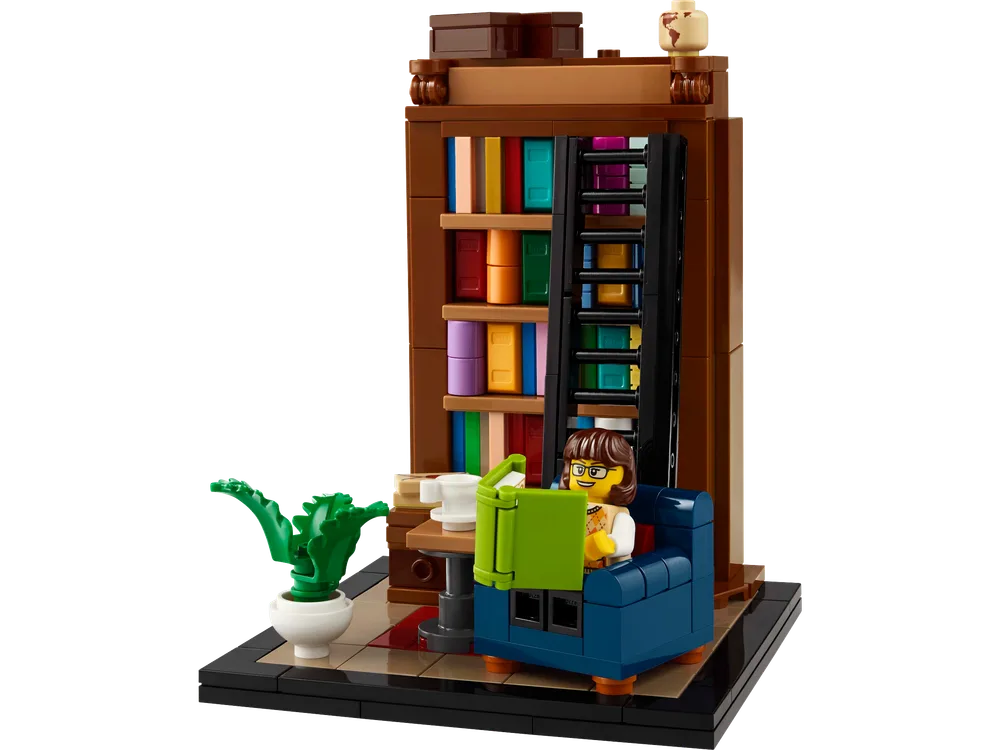 A LEGO minifigure reads a book in a blue armchair next to a tall, brown bookshelf filled with colorful books and a green potted plant on a black baseplate.