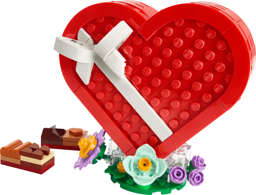 A fully assembled LEGO Creator 40759 Valentine's Day Heart Box, featuring a red heart-shaped box with a white ribbon, sitting on a clear and green floral base. Several brick-built chocolates are arranged next to it.