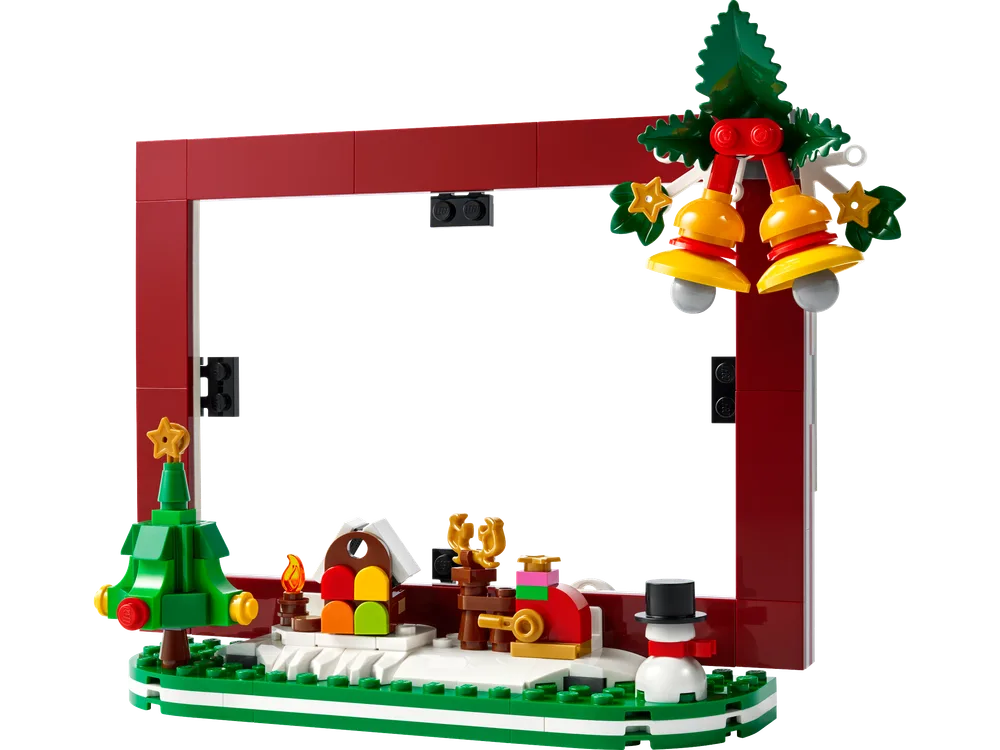A front, slightly angled view of the LEGO Christmas Picture Frame, set 40702, showcasing the red brick frame with a white interior for a photo. The base features a small winter scene with a green Christmas tree, a colorful house, a brown reindeer, and a snowman, all on a white and green base. Christmas bells hang from the top right corner.