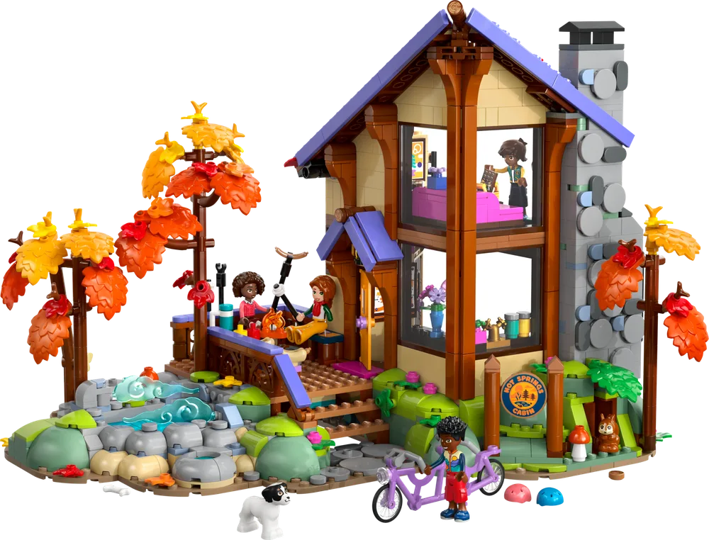 LEGO Friends Hot Springs Cabin set 42705, showcasing a two-story cabin with a purple roof, stone chimney, and autumnal trees. Two minifigures relax on the porch, a third is on a bicycle, and a dog plays nearby. A stream flows into a hot spring.