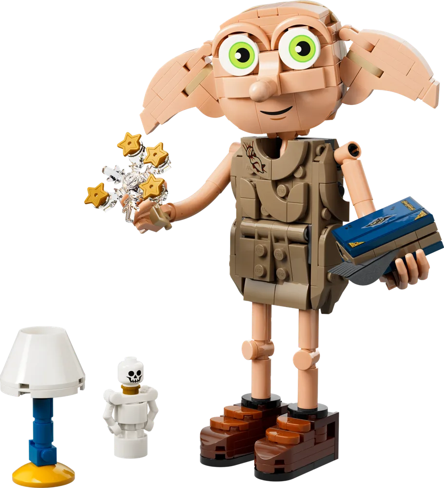 A LEGO brick-built Dobby the House-Elf stands on a white background, holding a shimmering burst of gold stars in one hand and Tom Riddle's diary in the other. A small white lamp with a blue base and a tiny white skeleton figure stand beside him.