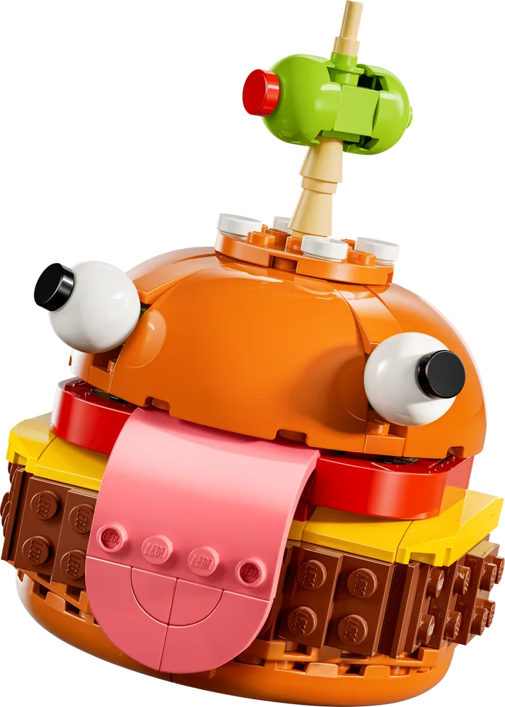 A close-up of the LEGO Fortnite Durrr Burger, featuring its orange bun, yellow cheese, brown patty, red tomato, and iconic pink tongue sticking out. Large white googly eyes are on either side of the bun, and a green olive with a red pimento is speared on top.