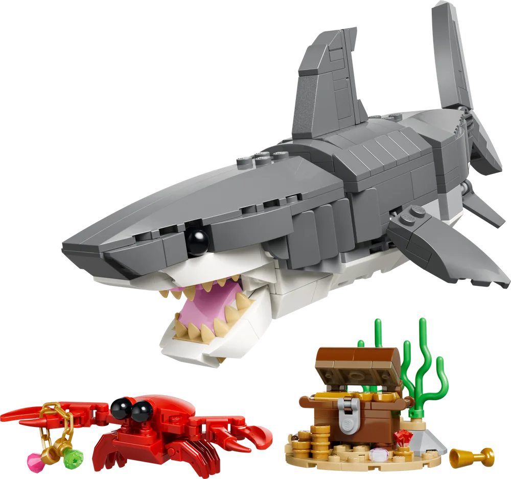 A large gray LEGO shark with an open mouth, showing teeth and pink gums, accompanied by a small red crab holding a charm and an open brown treasure chest filled with gold coins, on a white background.