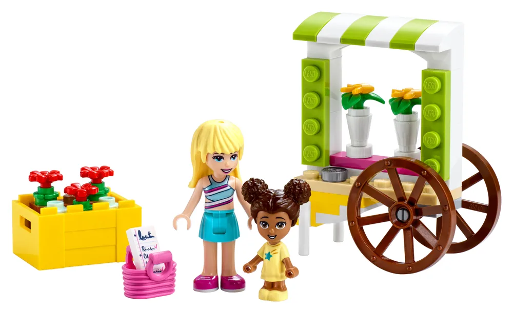 Two Lego Friends figures stand next to a white and green striped flower cart, a yellow planter box with red flowers, and a pink basket with a list.