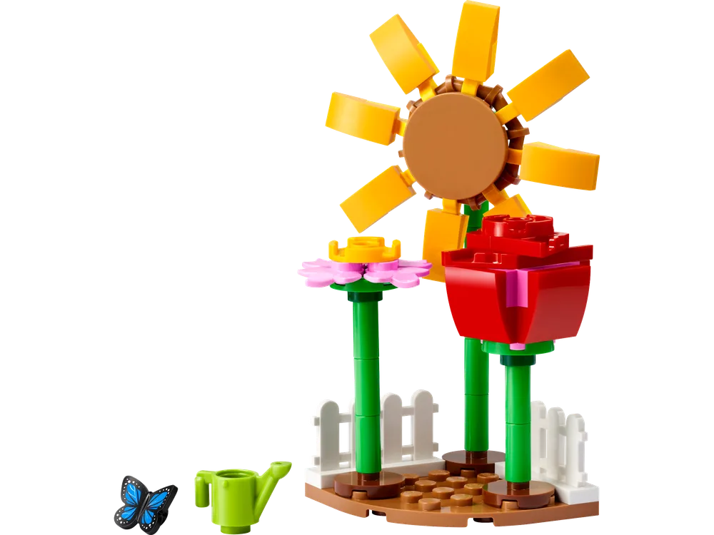 A built LEGO flower garden with a large yellow sunflower, a red rose, a green watering can, a blue butterfly, and a white picket fence on a brown base.