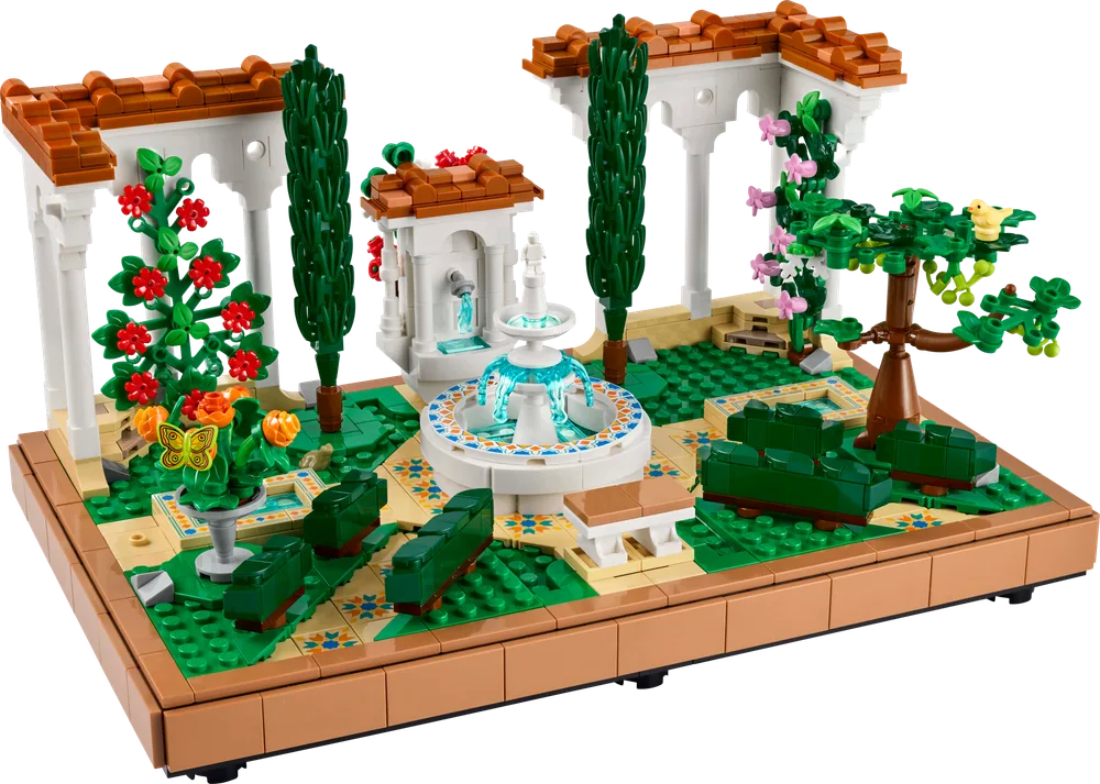 A wide, elevated view of the LEGO Icons Fountain Garden set, featuring a central tiered fountain, lush green foliage, white arcades with terracotta roofs, and decorative tiled pathways.