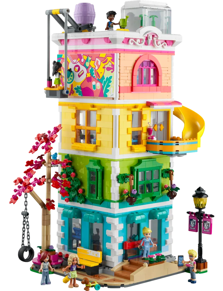 A vibrant, multi-story LEGO Friends Heartlake City Community Center model, viewed from the front, showcasing its four distinct colorful levels (teal, green, yellow, pink), a yellow slide, a working swing, a blossoming tree, and several minifigures enjoying various activities around the exterior.