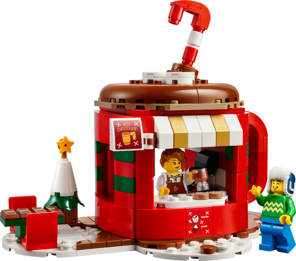 A vibrant red LEGO hot chocolate stand, shaped like a giant mug, with a chocolate-brown top and a red and white candy cane straw. A LEGO minifigure vendor serves hot chocolate from an interior counter, while a customer minifigure in a green sweater and earmuffs waves from the snowy base. A small LEGO Christmas tree and a bench with chocolate squares complete the festive scene against a white background.