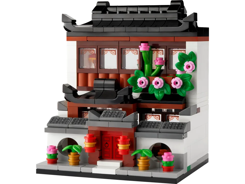 A LEGO model of a traditional East Asian house with a dark tiled roof, white walls, and dark brown accents. Pink flowers and green bamboo elements decorate the facade.