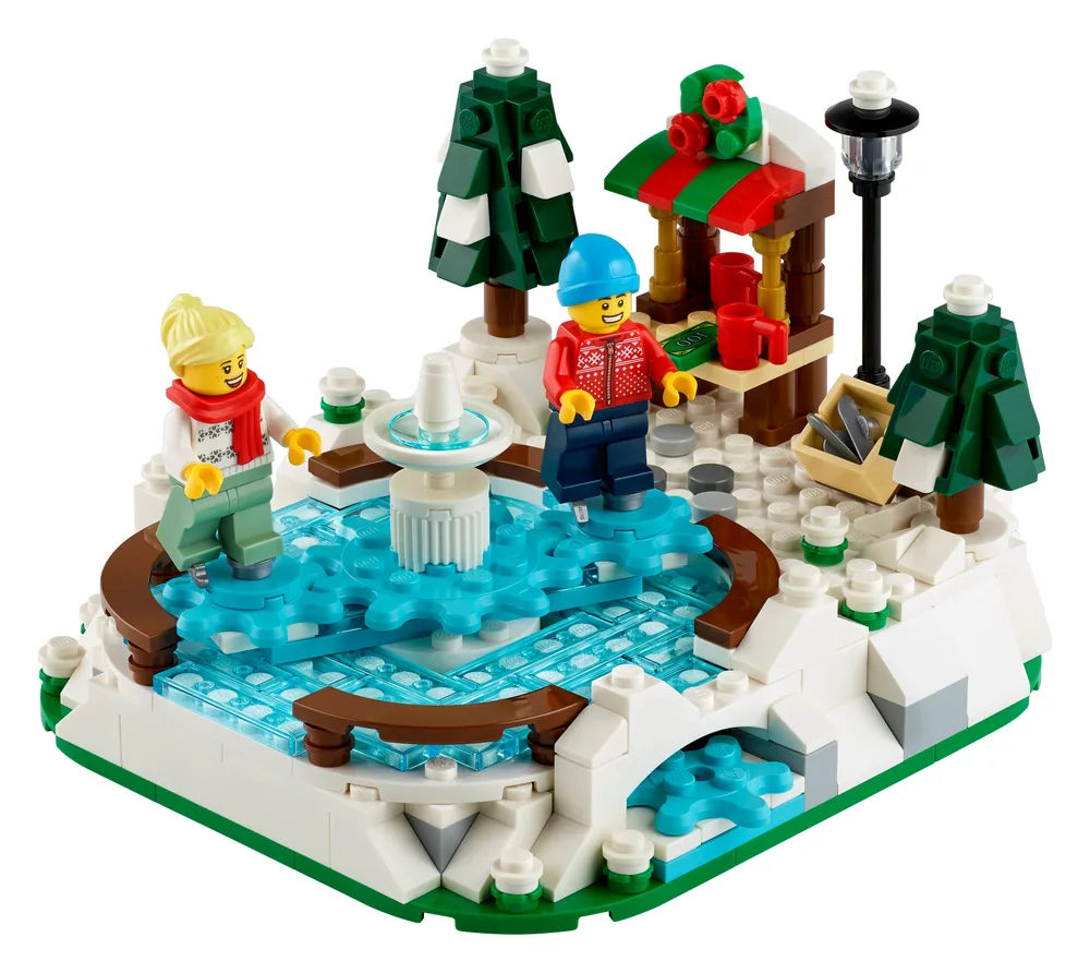 A close-up, slightly elevated view of the LEGO 40416 Winter Skating Scene set, featuring two minifigures, a rotating ice rink, a fountain, snow-covered ground, evergreen trees, a lamppost, and a small market stall.