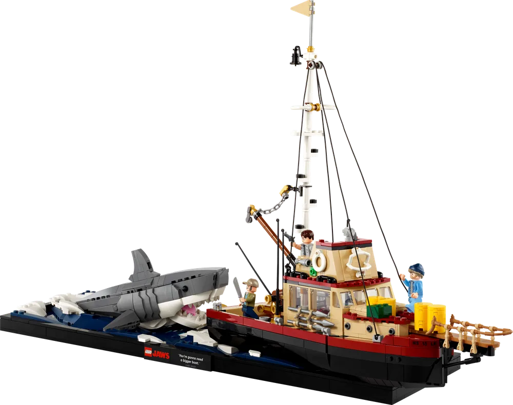 Full LEGO Ideas Jaws set showing the Orca boat in tan and dark red, battling a large grey and white brick-built shark. The boat is on a black display base with blue and white wave elements. Three minifigures are on the boat: Chief Brody, Quint, and Hooper.