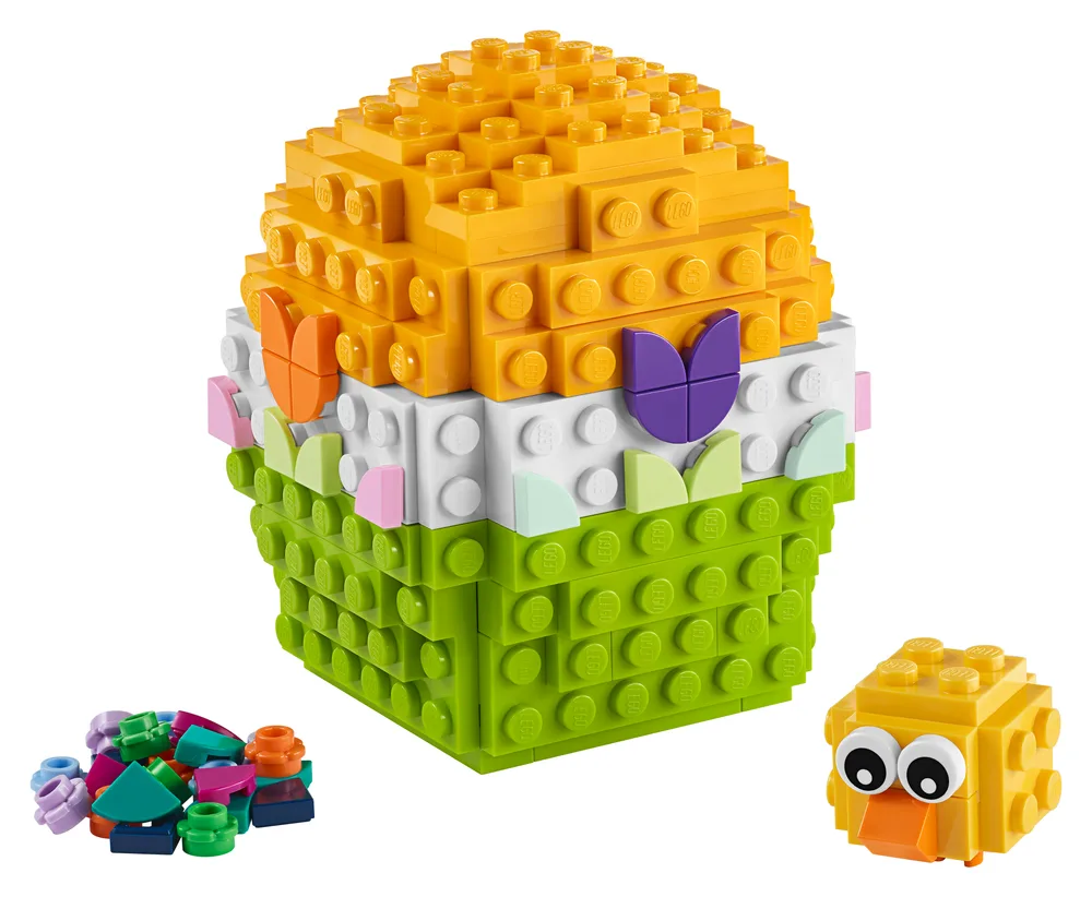 A LEGO brick-built Easter egg in yellow, white, and lime green with colorful floral decorations, accompanied by a small, blocky yellow chick figure and a pile of loose colorful LEGO parts.