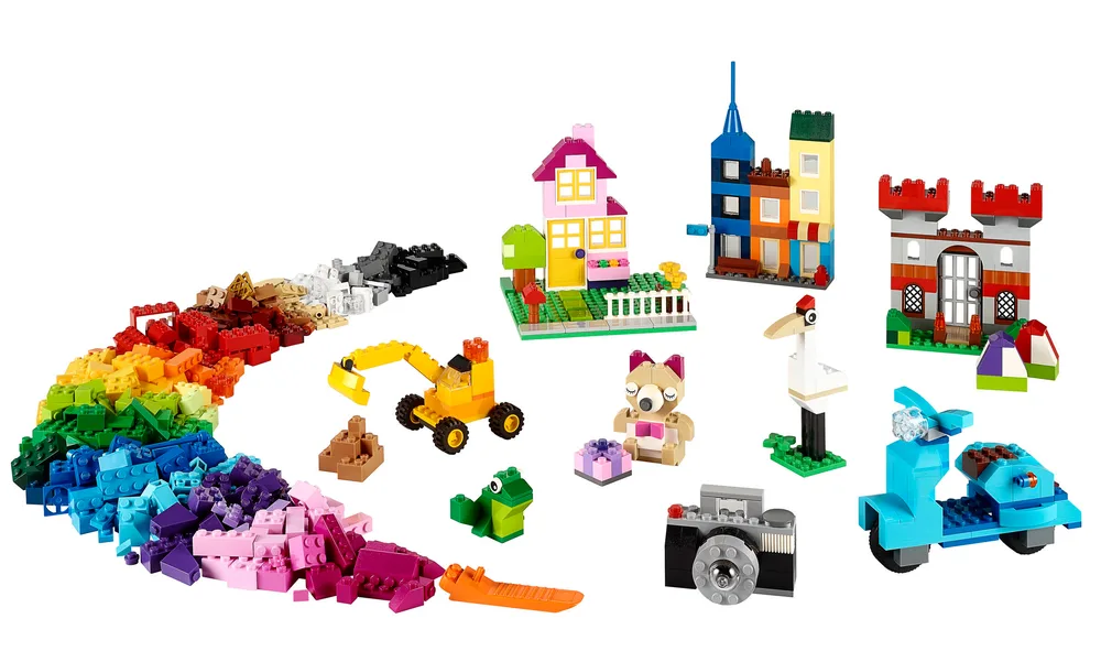 Assortment of LEGO Classic bricks in a rainbow arc with several completed models including a house, castle, scooter, excavator, and camera against a white background.
