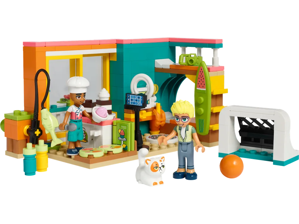 A LEGO Friends set showing a room with a kitchen area where Leo, wearing a chef's hat, is baking, and Olly is holding a phone. A cat sits nearby, and a surfboard and small soccer goal are also part of the scene.