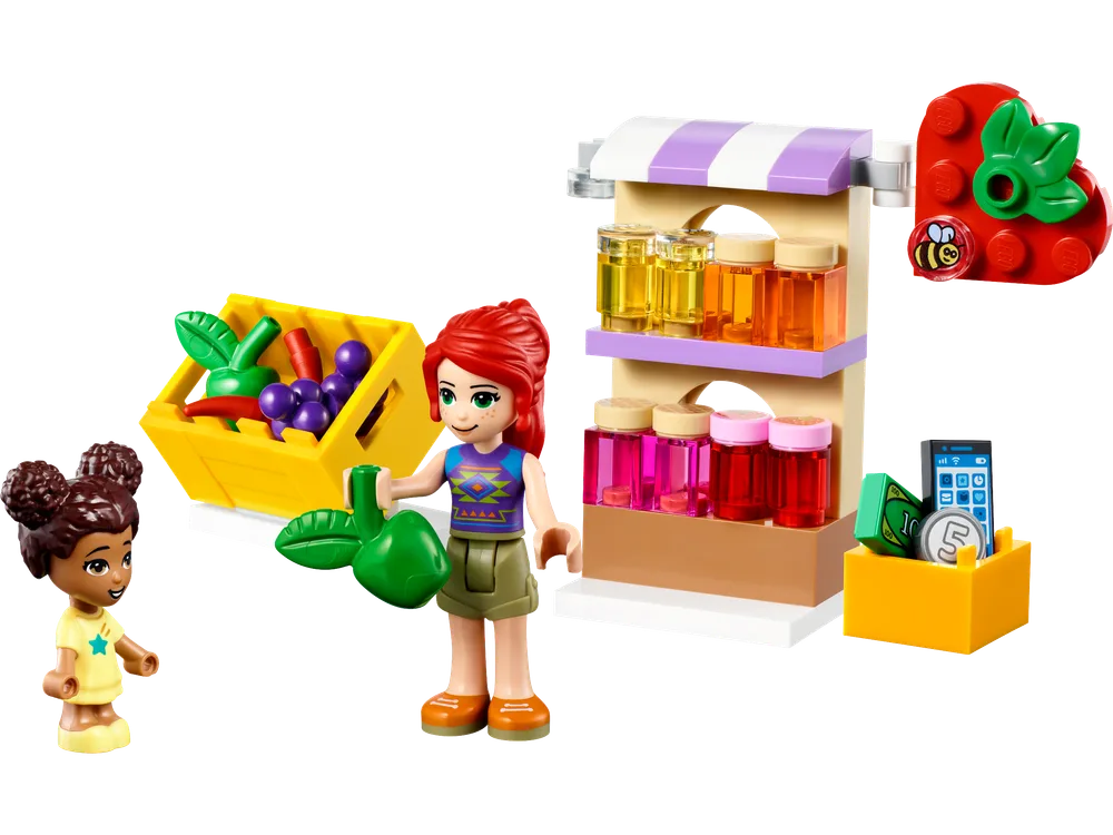 Two LEGO Friends minifigures, one adult and one child, stand next to a market stall displaying bottled liquids, a crate of fruit, and a cash register setup.