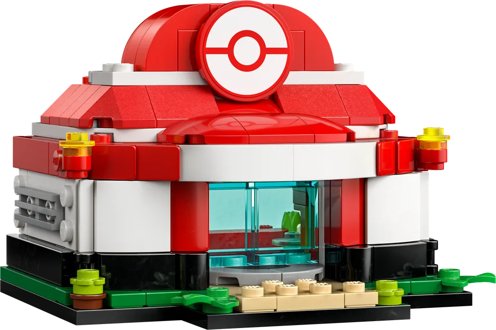 A LEGO build of a miniature Pokémon Center, featuring red domed roofs, white walls, and trans-clear blue windows, set on a green base with tan steps. A prominent red and white Poké Ball icon is on the front facade.