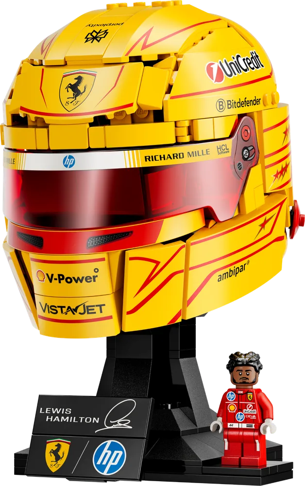 Lewis Hamilton's Ferrari F1 helmet built from LEGO bricks, featuring yellow and red colors, a clear visor, and sponsor logos on a black display stand. A Lewis Hamilton minifigure stands beside it.