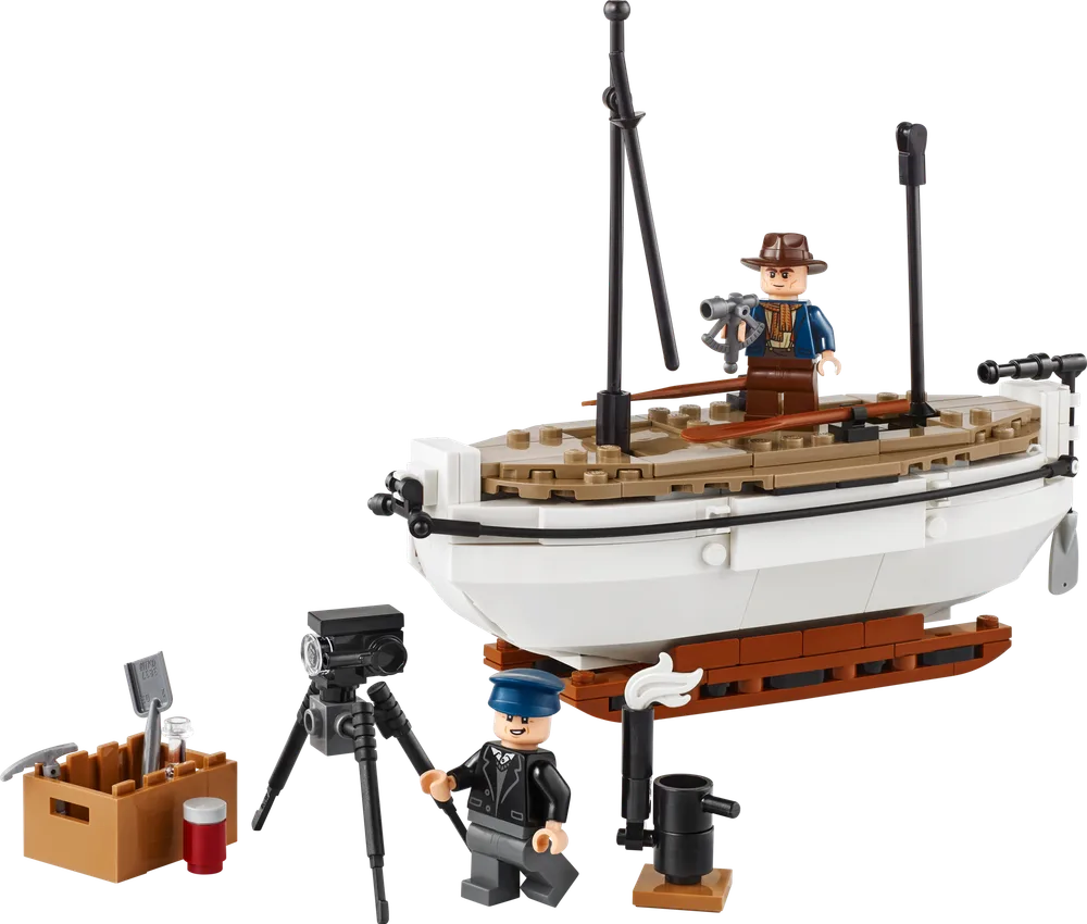 A LEGO set of Shackleton's lifeboat, featuring a white hull, tan deck, black mast, and two minifigures: one explorer with a sextant, and one cameraman with a tripod camera, alongside a wooden crate of tools and a lantern.