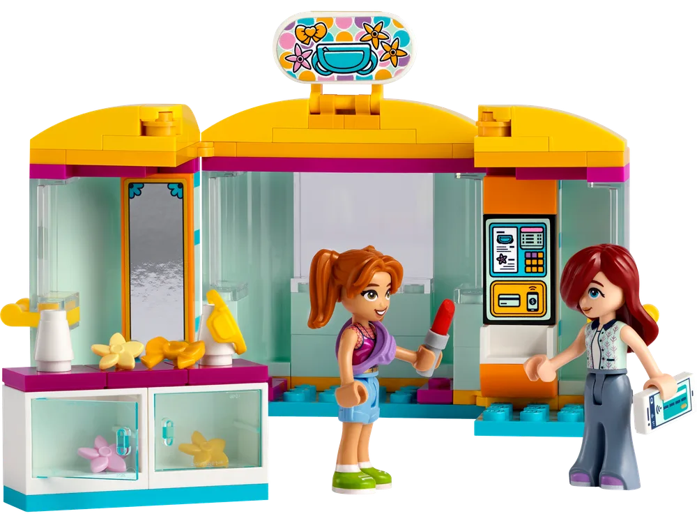 Two LEGO Friends minifigures, a red-haired shopper and a red-haired vendor, interact inside a brightly colored, open LEGO Friends accessory store. The store features a yellow roof, pink and blue accents, and displays of small accessories like lipstick and hair bows.