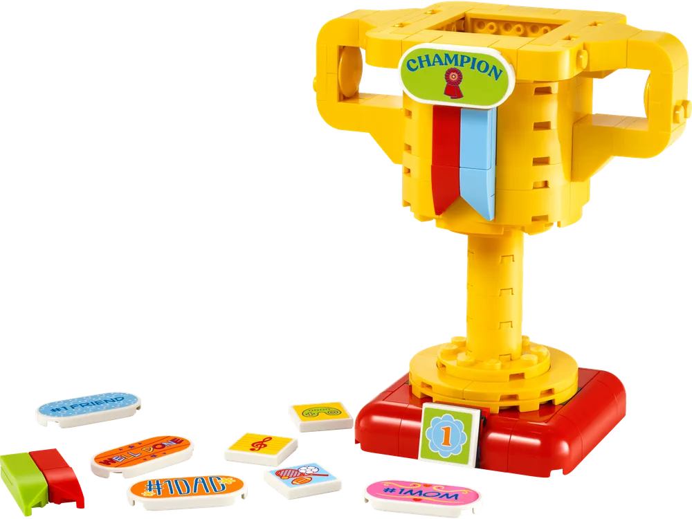 A disassembled yellow LEGO trophy with a red base and several customizable message tiles scattered around it, including '#1 Dad', '#1 Mom', and 'Welcome'.